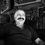 A cheerful man with a mustache smiles in a black and white portrait captured in a cozy setting in Mardin, Türkiye.