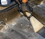 A hand in a glove using a vacuum to clean a car interior carpet, removing dirt effectively.